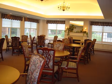 A well-lit dining room with multiple round wooden tables surrounded by upholstered chairs with floral patterns. Large windows with valances allow natural light to enter, and there is a fireplace with a mirror and greenery above it in the background.