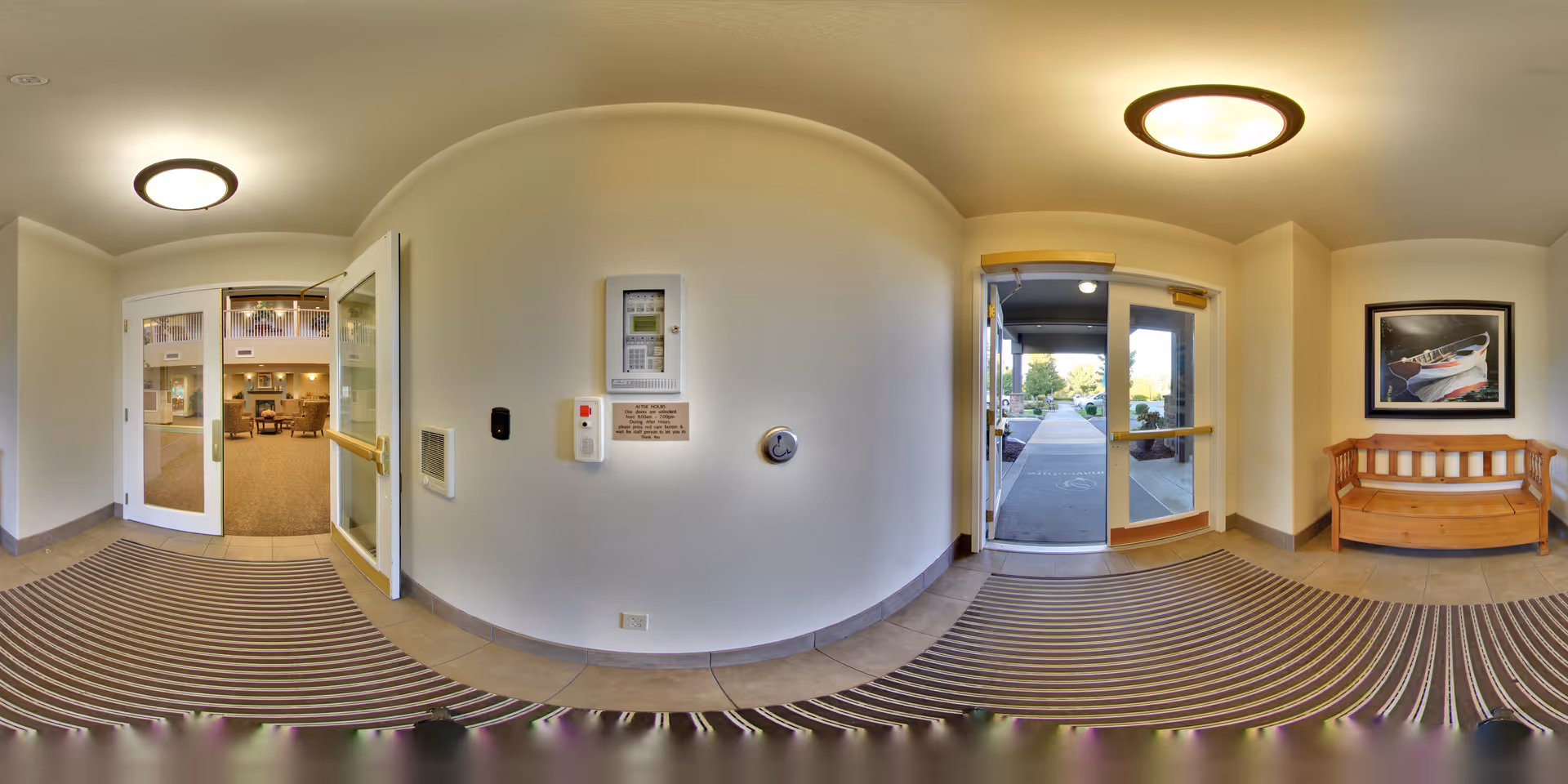 Entrance area of a senior living facility with double glass doors leading inside to a common area and another set of glass doors leading outside. The interior has beige walls, a striped floor mat, a wooden bench with a framed picture above it, and ceiling lights.