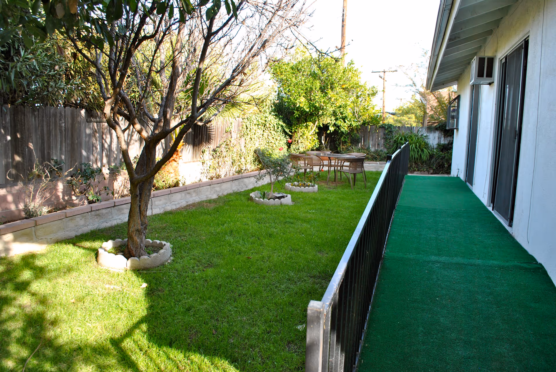Outdoor garden area with green grass, a tree surrounded by a circular stone border, and a small patio table with four chairs. The area is enclosed by a wooden fence and adjacent to a building with a green carpeted walkway and black railing.
