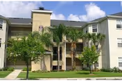 Front view of a three-story beige apartment building with balconies, palm trees, and a grassy lawn under a blue sky.