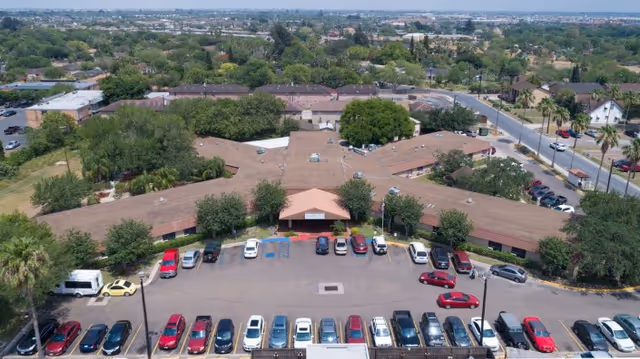 Aerial view of Alta Vista Rehabilitation & Healthcare facility showing a large, single-story building with a brown roof surrounded by trees and a parking lot filled with cars. The building is situated in a suburban area with roads and other buildings visible in the background.