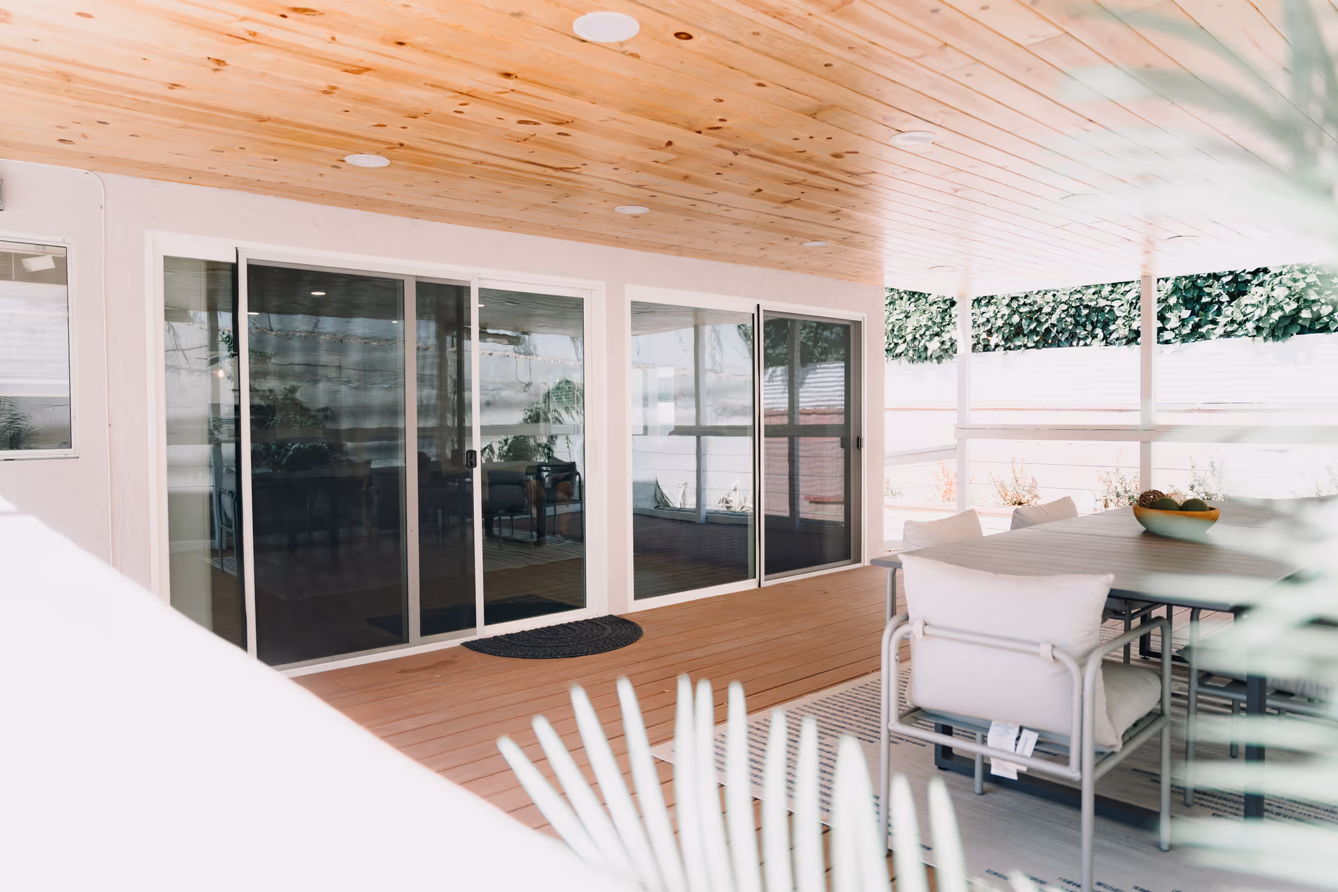 Covered outdoor patio area with wooden ceiling and floor, featuring a dining table with cushioned chairs and sliding glass doors leading inside. Greenery and a white fence are visible in the background.
