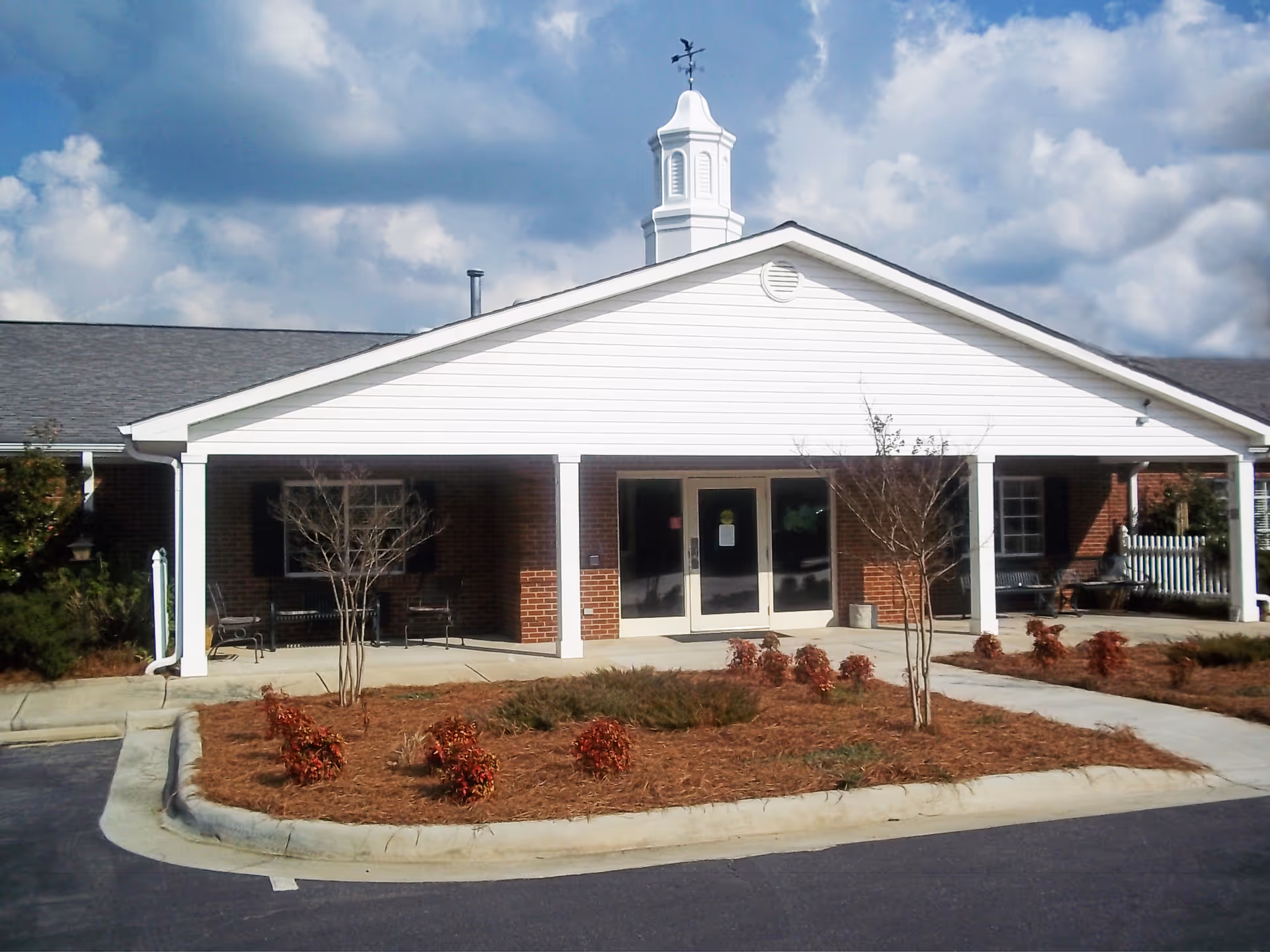 Front exterior view of a single-story brick building with a white covered entrance supported by columns, small landscaped bushes, and a weather vane on top of the roof under a partly cloudy sky.