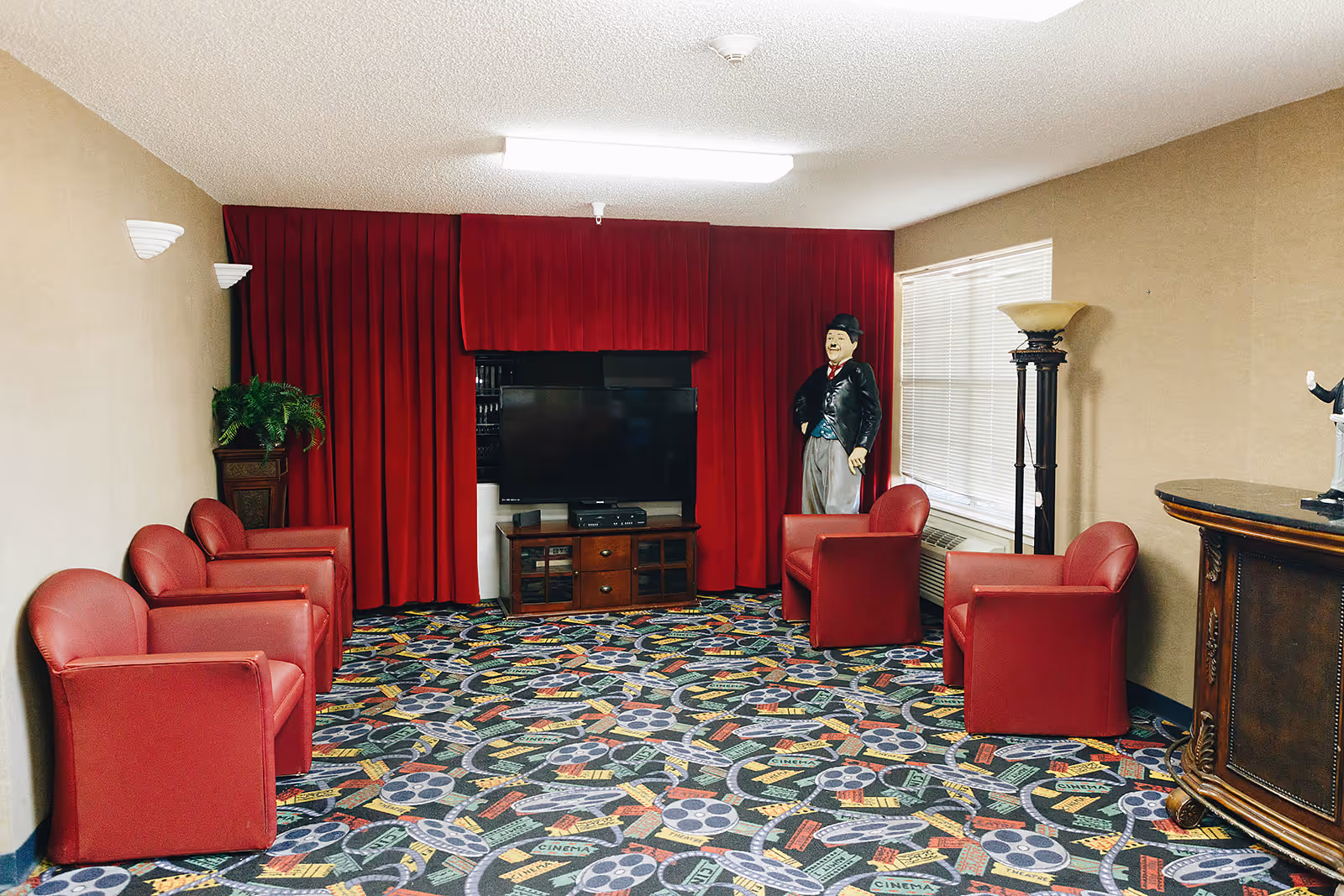 A lounge area with six red armchairs arranged around a television set on a wooden stand. The room has a colorful carpet with a cinema theme, beige walls, a window with blinds, a tall floor lamp, and a life-sized statue of a man in a suit and hat standing near the window. Red curtains cover the wall behind the television.
