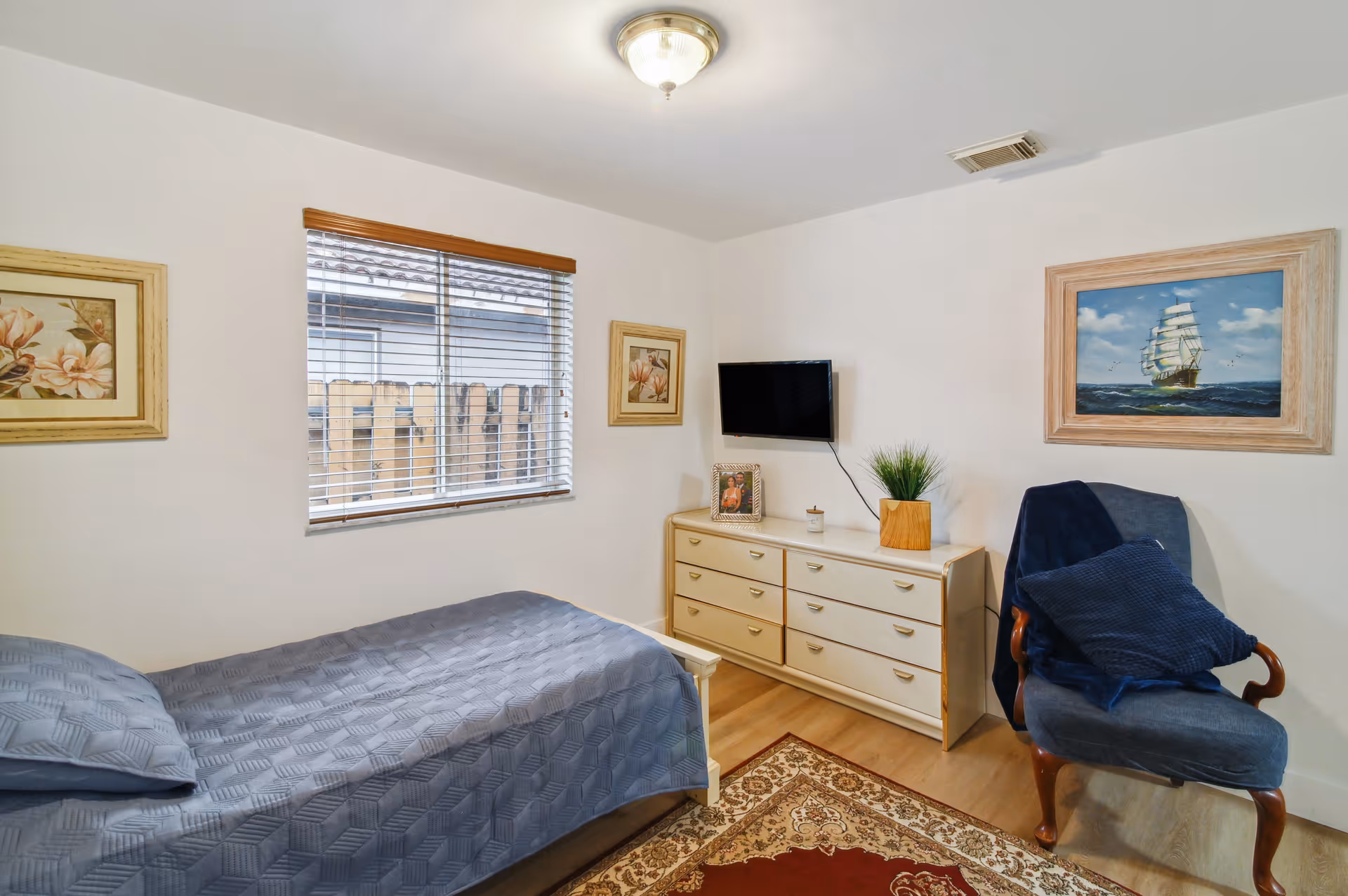A small bedroom with a single bed covered in a gray quilt, a window with blinds, a dresser with a plant and framed photo on top, a wall-mounted TV, a blue upholstered chair with a dark blue pillow and blanket, and framed artwork on the walls.