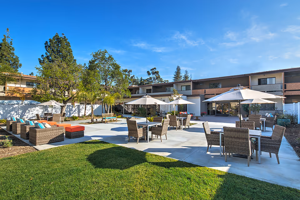 Outdoor patio area at Canyon Trails at Topanga Senior Living featuring multiple wicker chairs and tables with umbrellas, cushioned seating, green grass, trees, and a two-story building in the background under a clear blue sky.