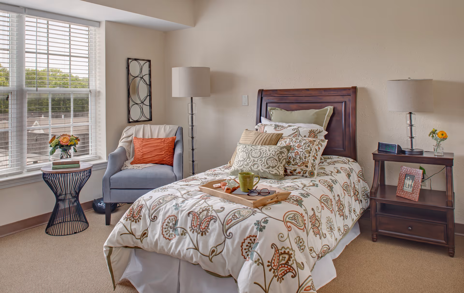 A cozy bedroom with a single bed featuring a floral patterned comforter and multiple pillows. A wooden headboard is behind the bed. On the bed is a wooden tray holding a green mug, a pair of glasses, and a napkin. To the right of the bed is a wooden nightstand with a lamp, a digital clock, a framed photo, and a small vase with a yellow flower. To the left of the bed is a gray armchair with an orange pillow and a light throw blanket. Next to the chair is a round side table with a vase of flowers and two books. A large window with white blinds lets in natural light, and a decorative wall mirror hangs on the wall behind the chair.