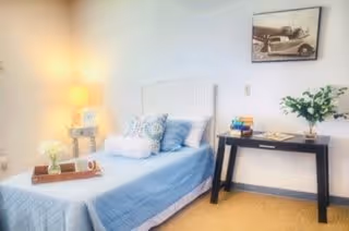 A bright and cozy bedroom with a single bed dressed in light blue bedding and multiple pillows. A small wooden nightstand with a lamp and decorative items is placed next to the bed. Across from the bed is a black console table with books and a vase of greenery. A framed vintage car photograph hangs on the white wall above the table.