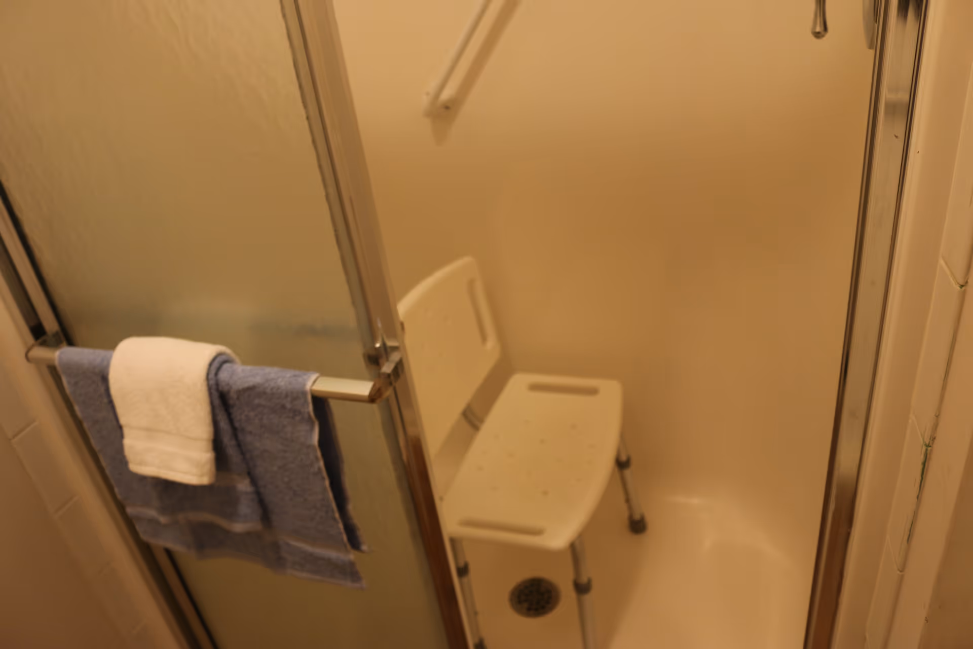 A shower stall with a white plastic shower chair inside. The shower has a frosted glass sliding door with a towel bar holding a blue towel and a white washcloth. There is a grab bar mounted on the wall inside the shower.