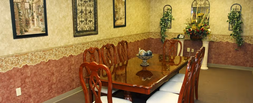 Long polished wooden dining table with upholstered chairs in a decorated dining room with framed art and plants.