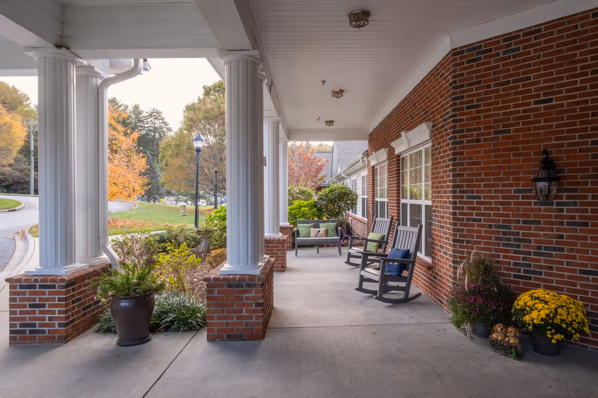 Covered outdoor porch area with white columns and brick walls, featuring rocking chairs with cushions, a bench with pillows, potted plants, and a welcome sign. Trees and a street are visible in the background.