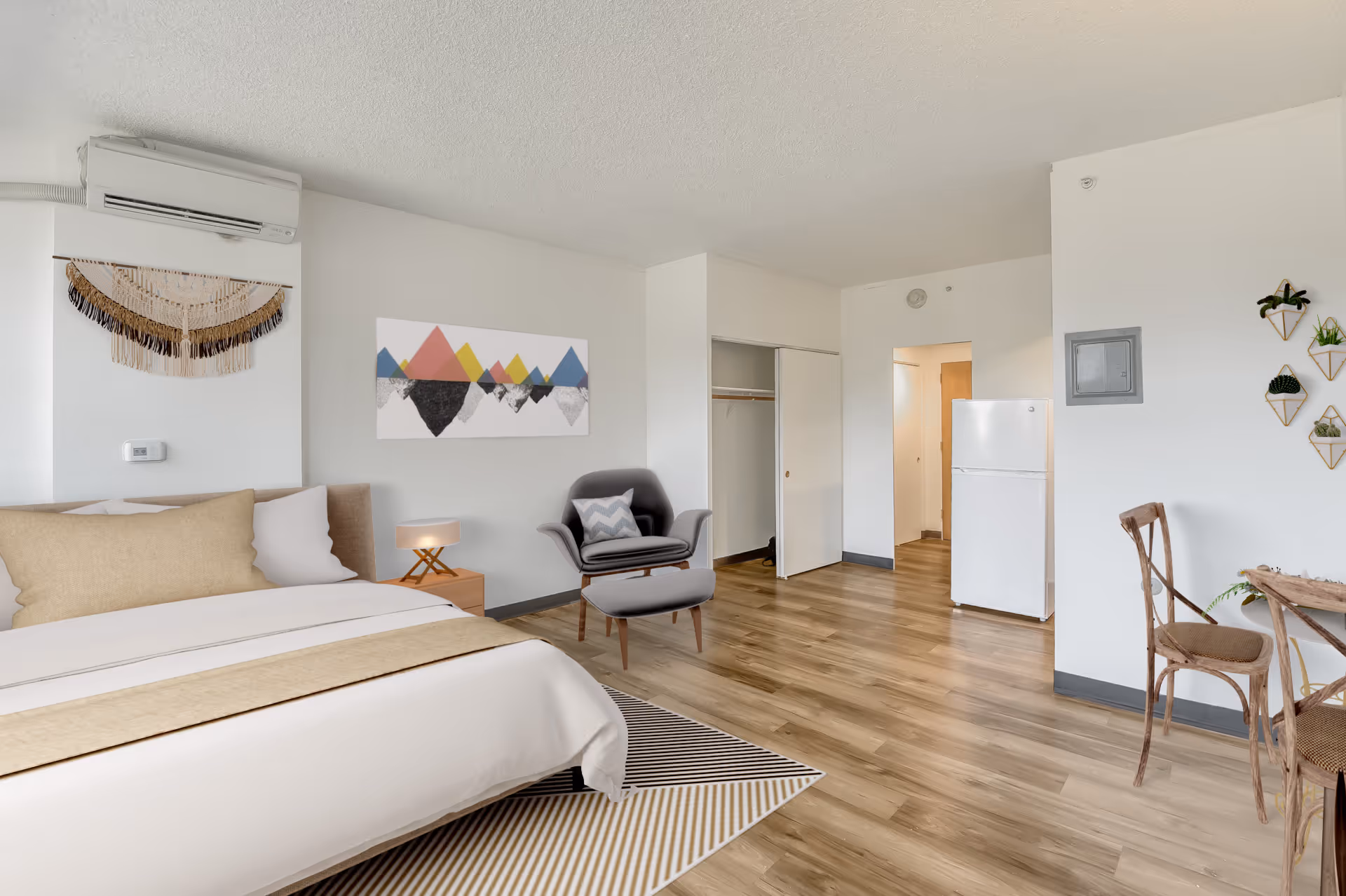 Bright studio-style bedroom with a made bed, armchair and ottoman, small dining table, and a refrigerator against white walls.