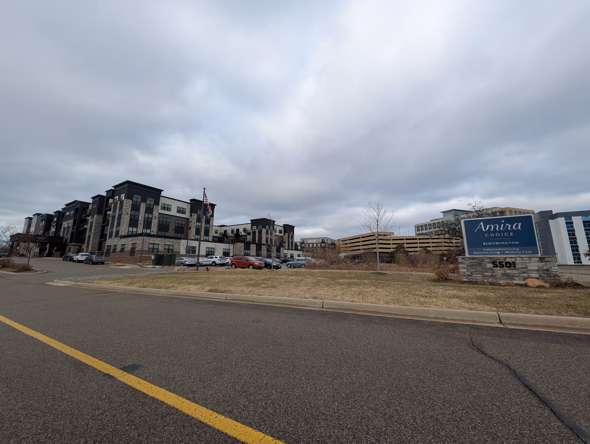 Exterior view of Amira Choice Bloomington facility showing a multi-story building with a parking lot in front, an American flag on a pole, and a large sign displaying the facility name and services including Independent Living, Assisted Living, and Memory Care. The sky is cloudy and the surrounding area includes some grass and trees.