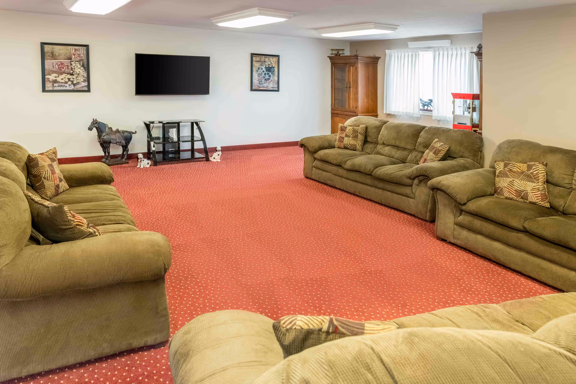 A spacious living room with red carpet flooring and three green upholstered sofas arranged around the room. The walls are white with two framed pictures and a flat-screen TV mounted on one wall. There is a wooden cabinet near a window with white curtains, and decorative items including a horse statue and small dog figurines are placed around the room.