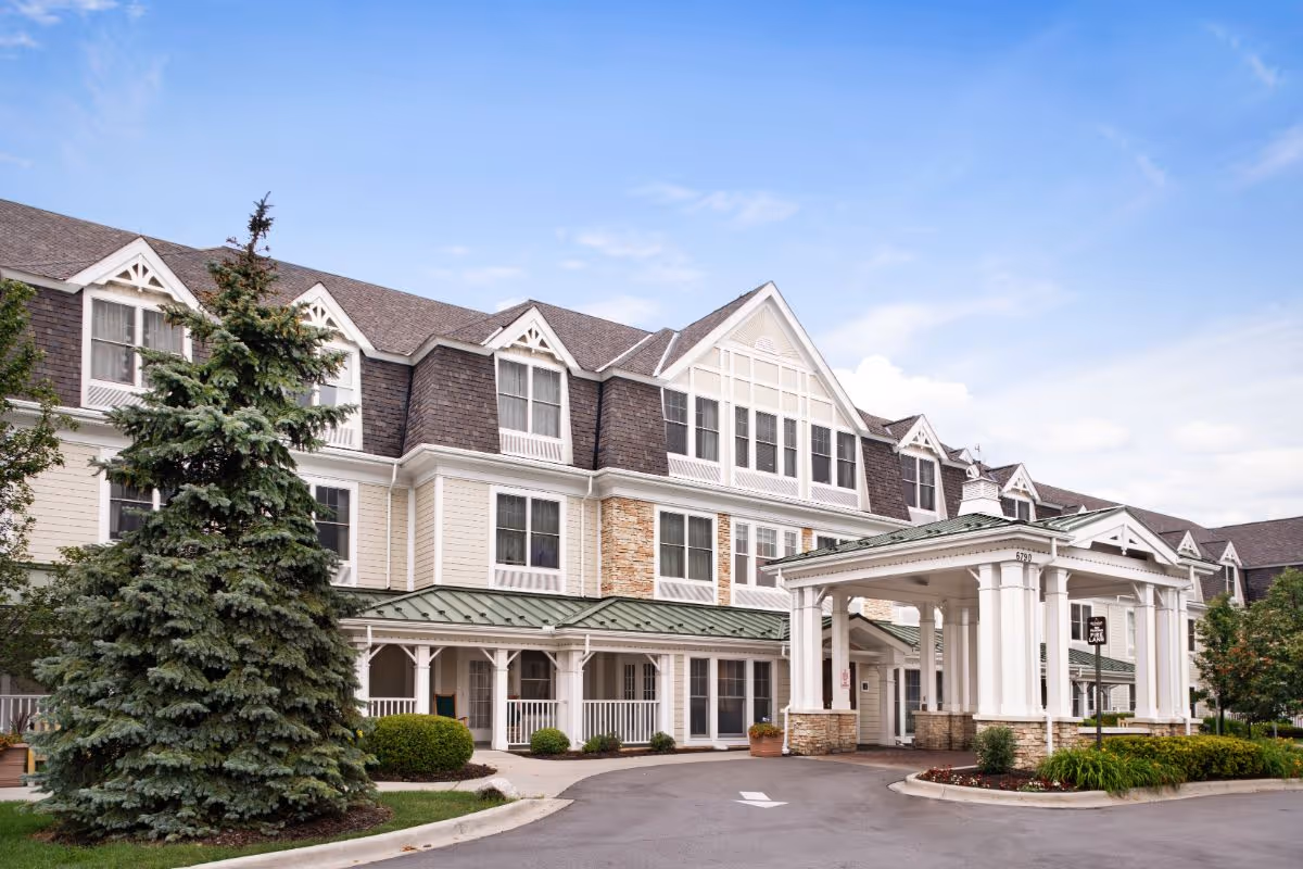 Front exterior of a multi-story senior living facility with a covered porte-cochère, dormer windows, and landscaped driveway.