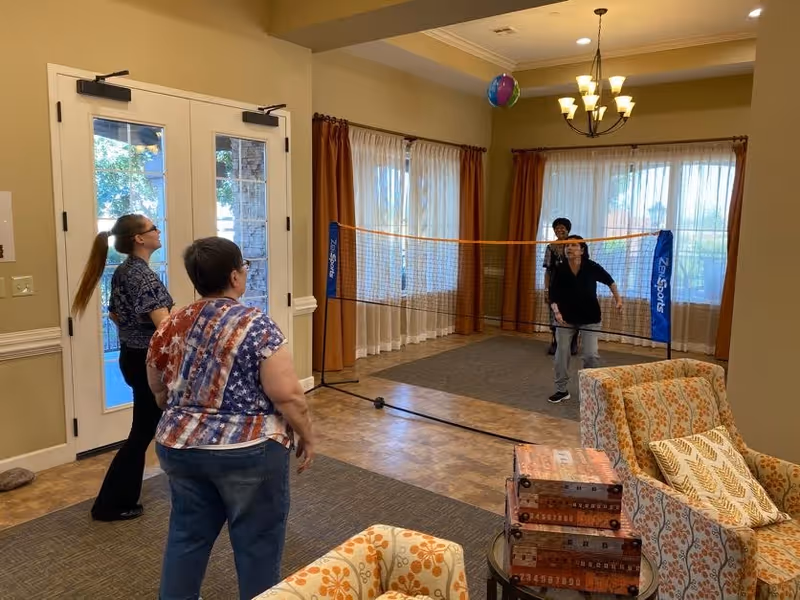 Residents playing a net game in a senior living community lounge with chairs, large windows, and double doors.