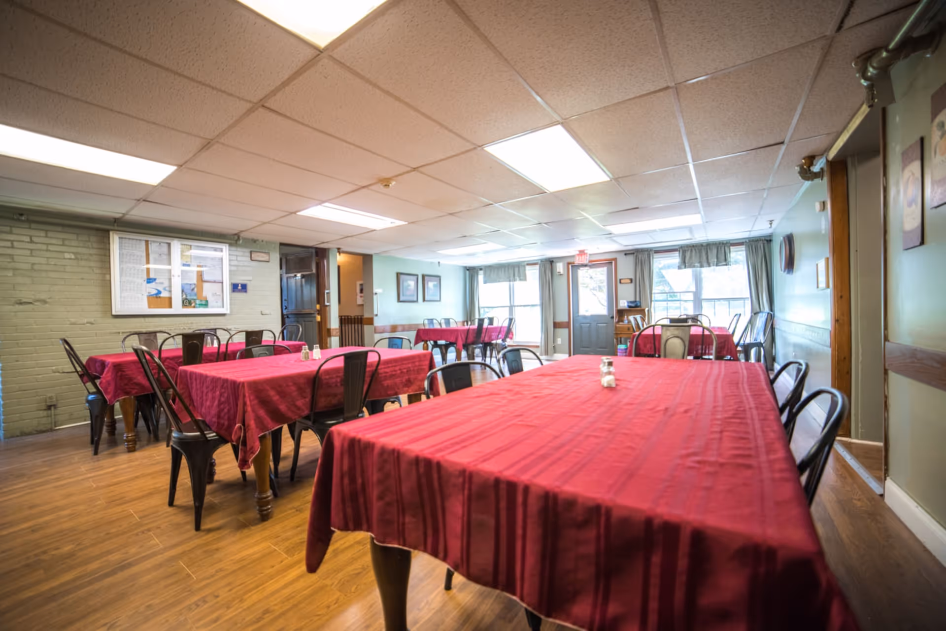 Communal dining room with multiple tables covered in red tablecloths and metal chairs under fluorescent lighting.