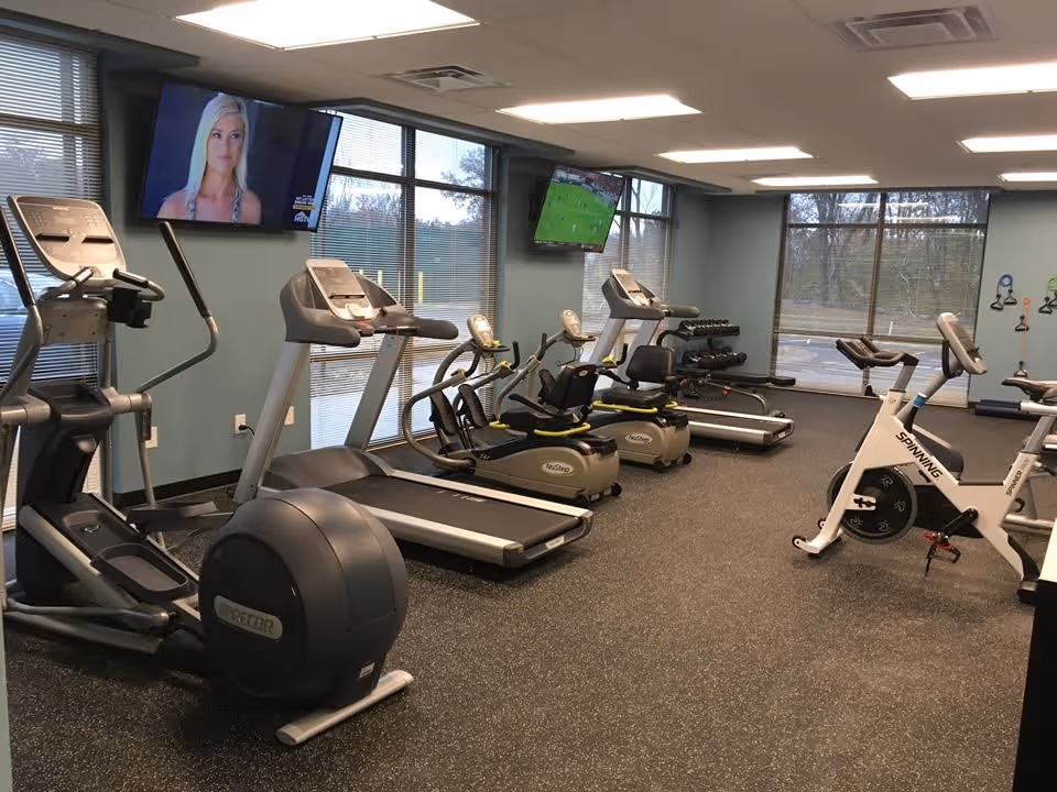 Interior view of a fitness room with exercise equipment including treadmills, stationary bikes, elliptical machines, and weight racks. Two wall-mounted televisions are visible, and large windows provide natural light.