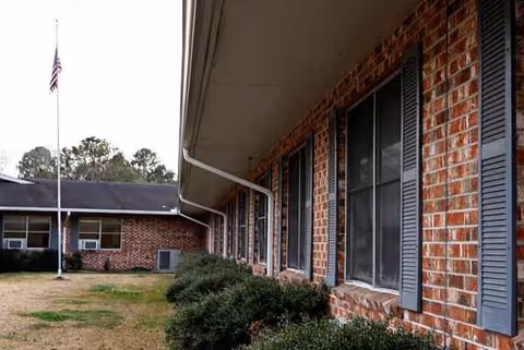 Brick senior living building exterior with windows, shutters, flagpole, and trimmed hedges.