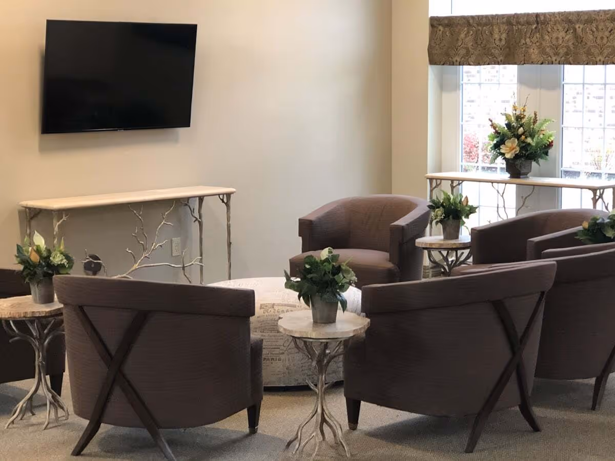A cozy seating area with four brown upholstered armchairs arranged around a round ottoman with script print. There are small round side tables with floral arrangements on them. A flat-screen TV is mounted on the wall above a decorative console table with a branch-like design. Large windows with patterned valances allow natural light into the room.