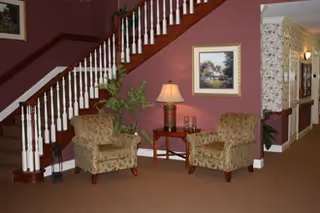 Lobby seating area with two upholstered chairs, a small table and lamp beneath a staircase.