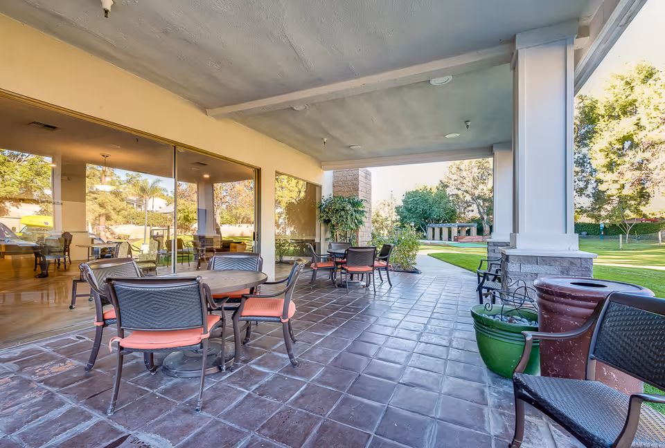 Covered outdoor patio area with several round tables and chairs with orange cushions, large windows reflecting the interior dining area, and a view of a green lawn and trees in the background.