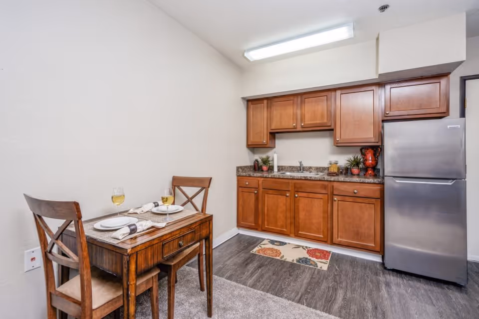 Small kitchen area with wooden cabinets, a stainless steel refrigerator, and a countertop with a sink. Next to the kitchen is a small wooden dining table set for two with plates, napkins, and glasses of white wine. The floor is a combination of carpet and wood-like flooring, and the walls are painted white.