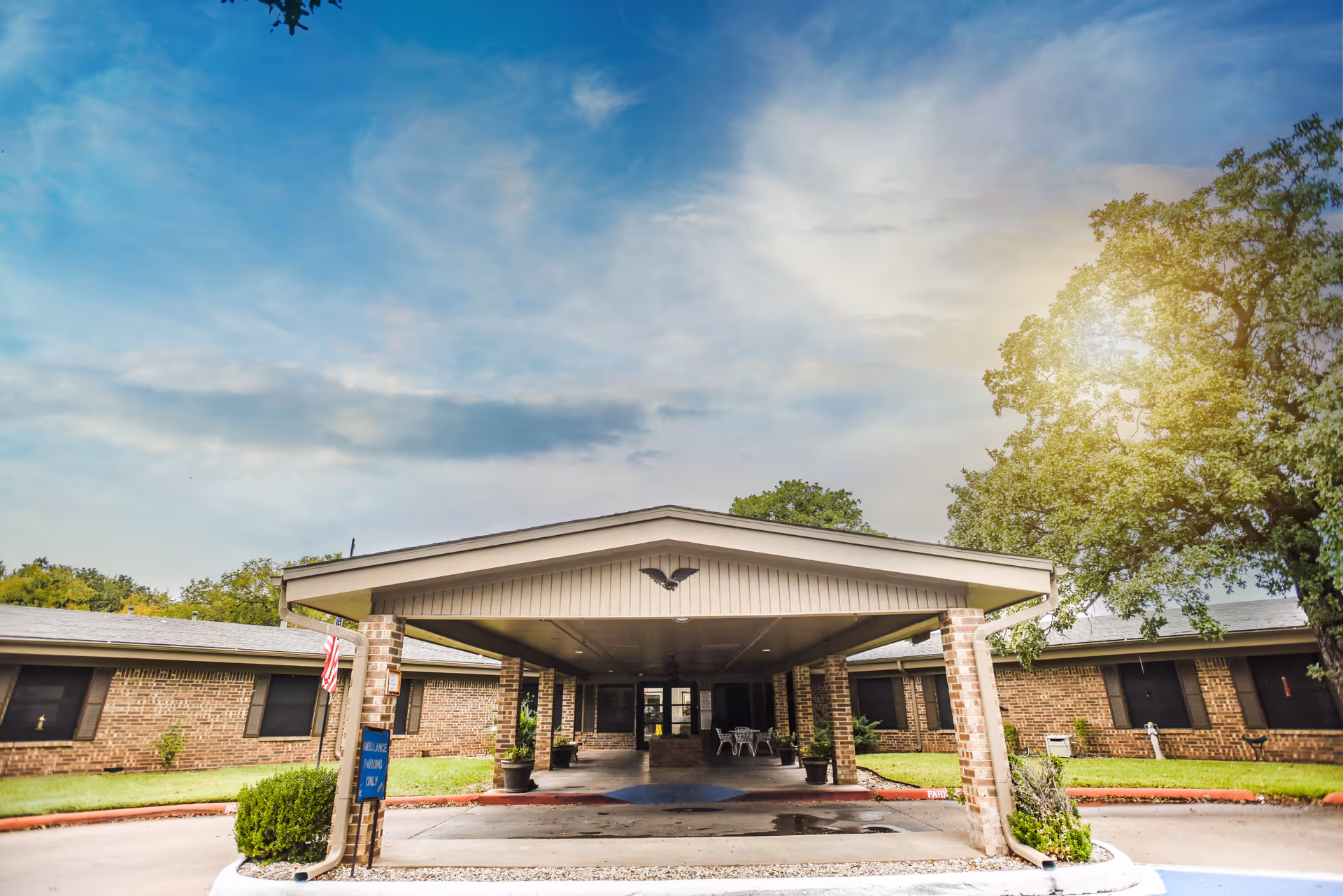 Front entrance of a single-story brick care center with a covered porte-cochere, flag, and surrounding trees under a blue sky.