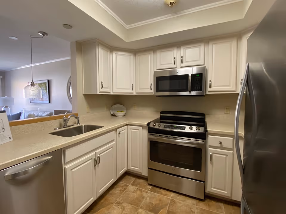 A modern kitchen with white cabinets, a stainless steel dishwasher, oven, microwave, and refrigerator. The kitchen has a beige countertop and tiled floor. There is a sink with a faucet and a bowl of green apples on the counter. The kitchen opens to a living area with a hanging light fixture and framed artwork on the wall.