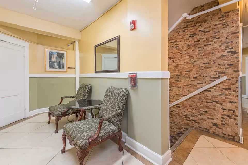 A small seating area in a hallway with two upholstered armchairs and a glass-top table between them. The walls are painted yellow and green with white trim, and there is a large mirror and a framed picture on the walls. To the right, there is a staircase with a stone-textured wall and a handrail.