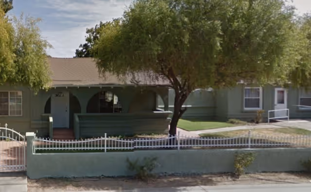 Exterior view of a single-story building with a light green facade, a small front porch with arches, a white metal fence, and trees providing shade in front of the building.