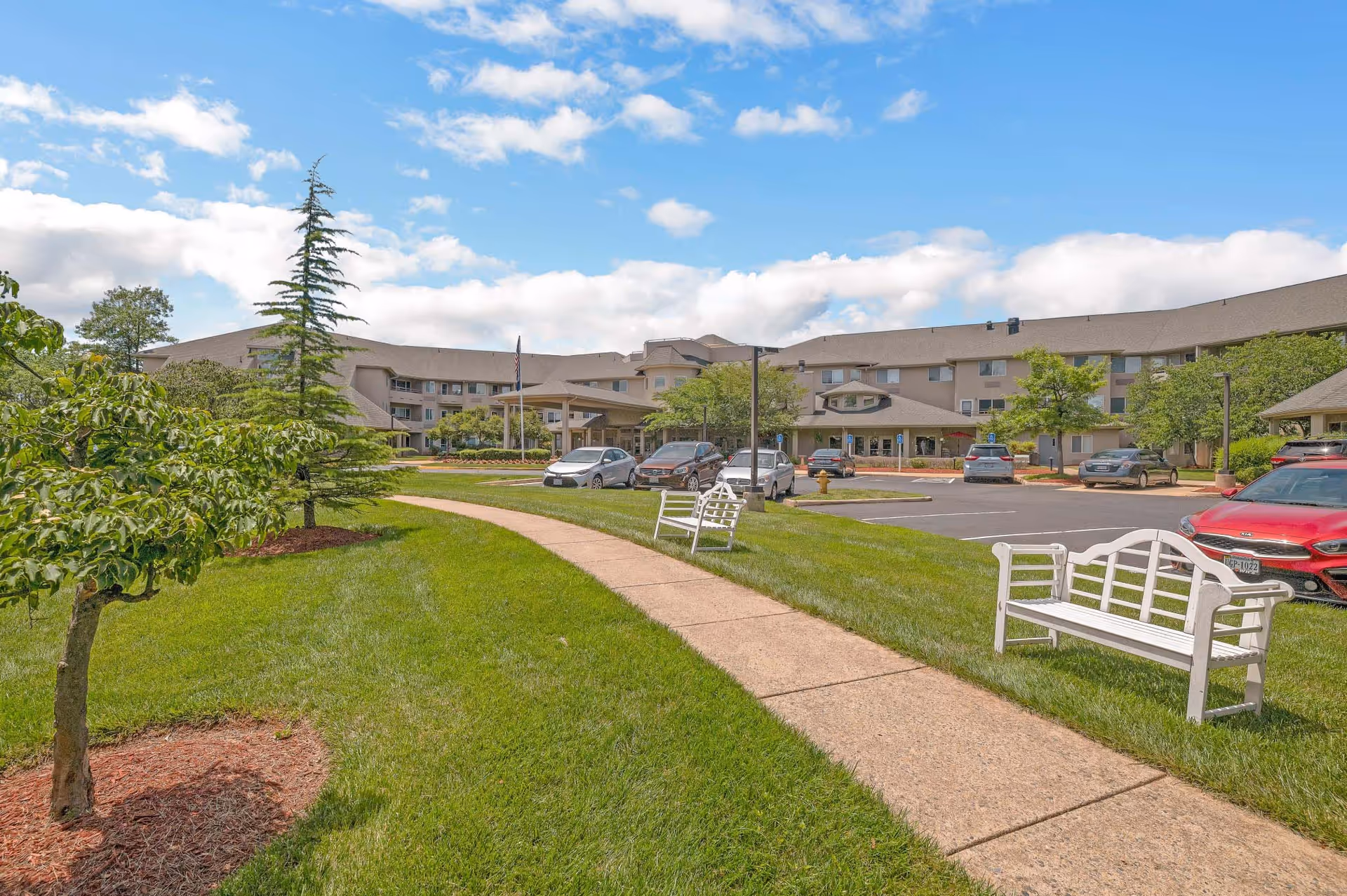 Outdoor view of The Fairmont by Barclay House senior living facility showing a curved sidewalk flanked by green grass and white benches, with trees and a parking lot in front of a large multi-story building under a partly cloudy blue sky.