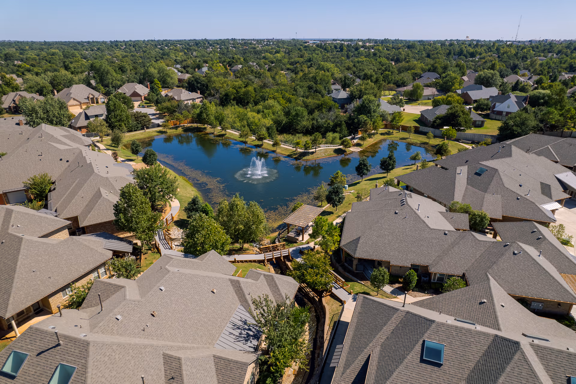 Aerial view of a senior living facility named Mon Abri featuring multiple buildings with gray roofs surrounding a central pond with a water fountain. The area is landscaped with trees, walking paths, and benches, with a residential neighborhood visible in the background under a clear blue sky.