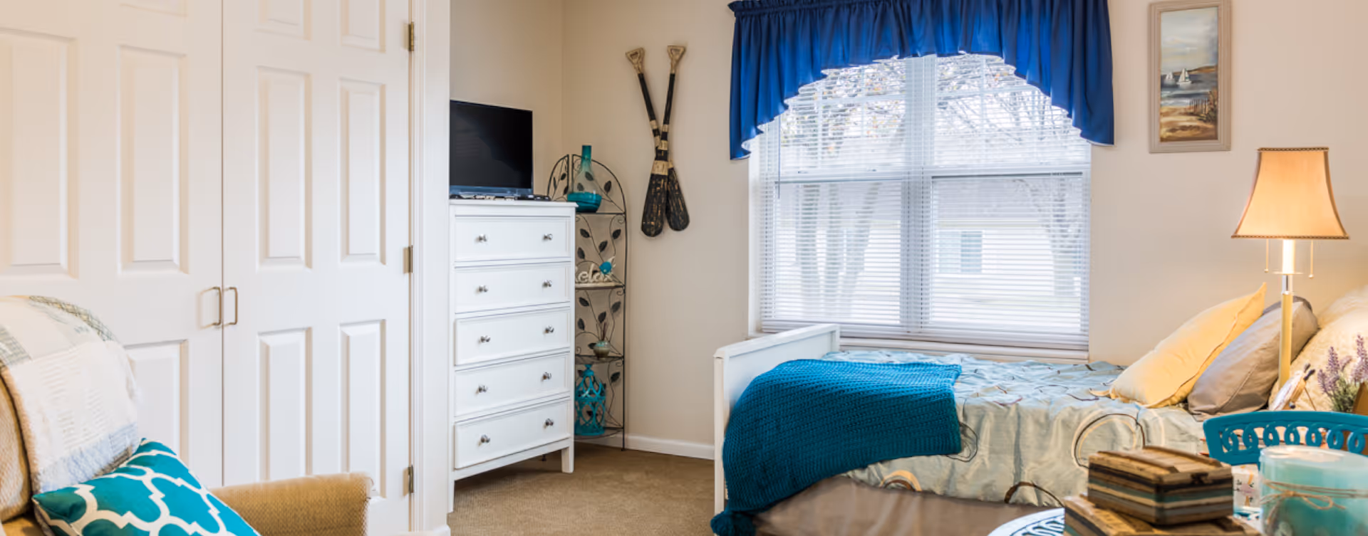 A bright, furnished bedroom with a bed under a window, white dresser, armchair, and lamp with teal accents.
