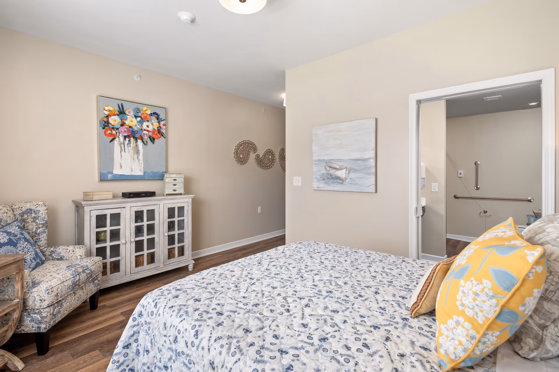 A cozy bedroom in a senior living facility featuring a bed with floral-patterned bedding and yellow accent pillows. There is a patterned armchair with a blue cushion, a white cabinet with glass doors, and two paintings on the beige walls. The room has wood flooring and an open doorway leading to a bathroom with grab bars.