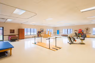 A spacious physical therapy or rehabilitation room with large windows letting in natural light. The room contains parallel bars for walking exercises, exercise equipment including a treadmill and stationary bike, therapy tables, and various therapy tools and supplies along the walls.