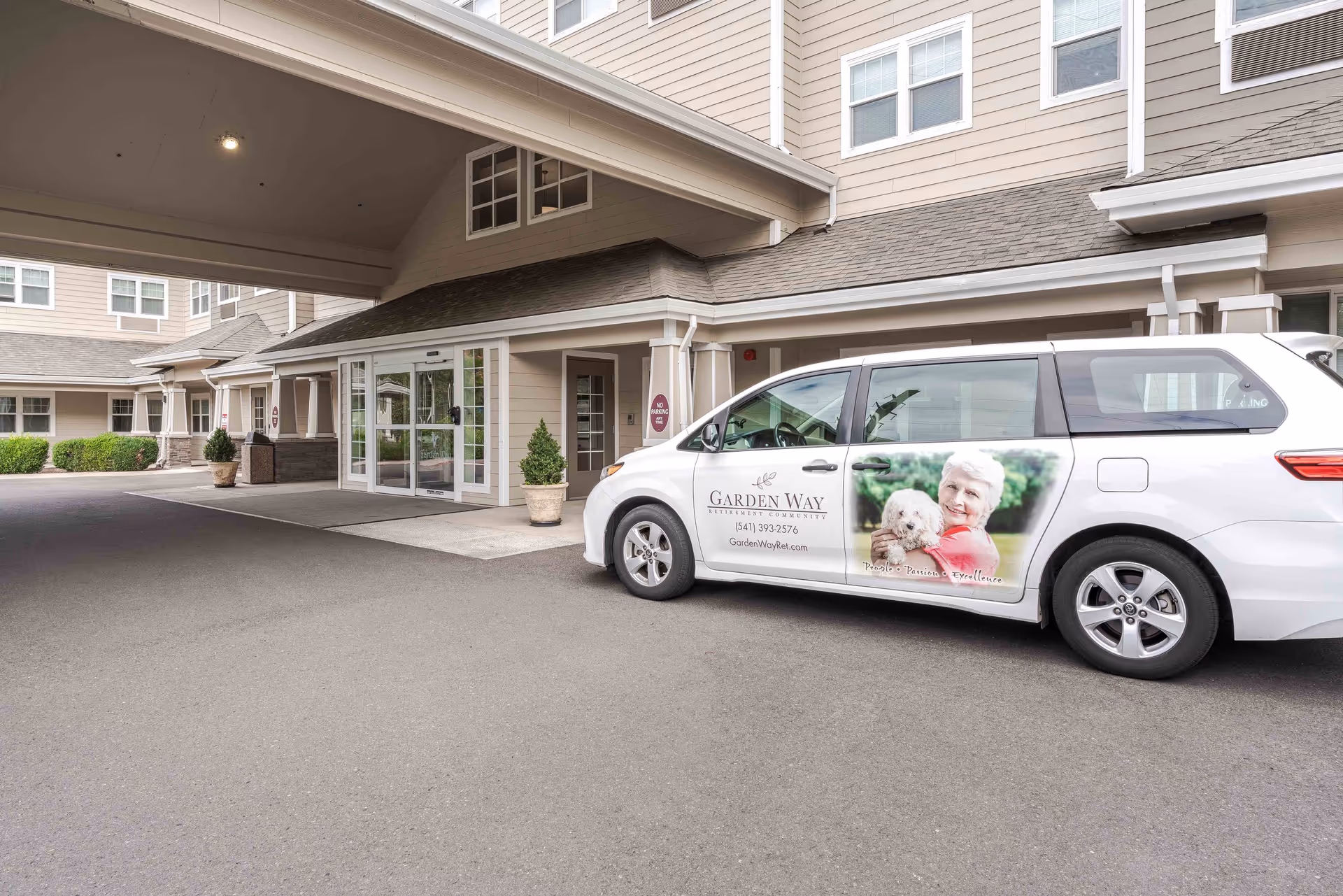 Entrance of Garden Way Retirement Community with a covered driveway and a white van parked nearby. The van features an image of an elderly woman holding a small dog and the community's contact information on its side.