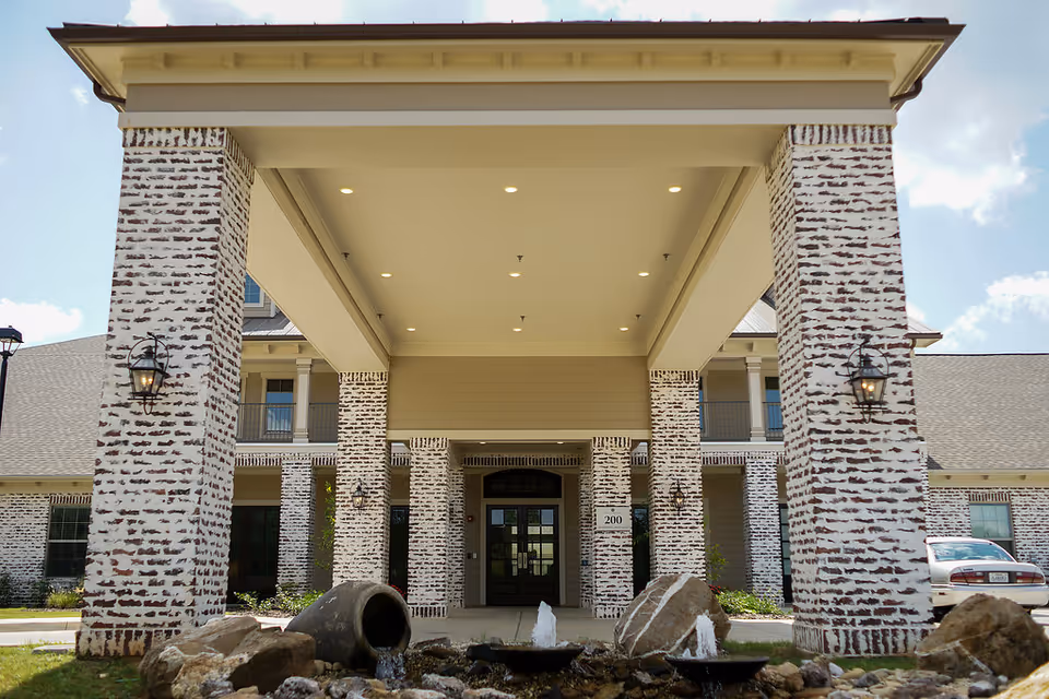 Covered front entrance of an assisted living building with whitewashed brick columns and a small fountain in the foreground.