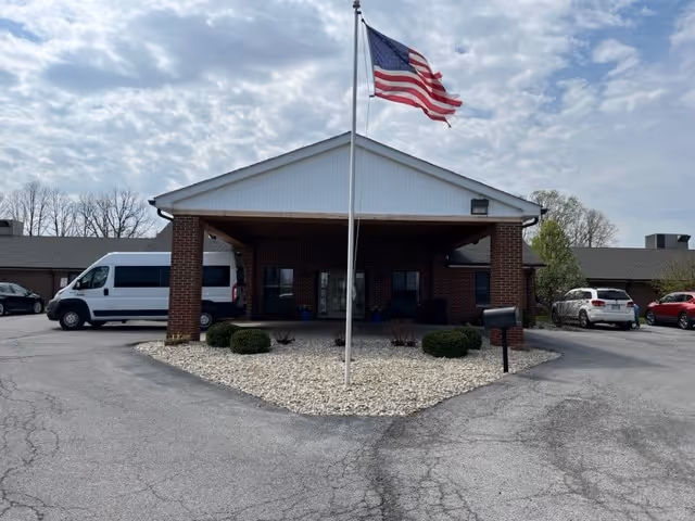 Front exterior view of Kenton Nursing & Rehabilitation Center showing a brick building with a covered entrance, an American flag on a flagpole in the center, and several parked vehicles including a white van and cars. The sky is partly cloudy.