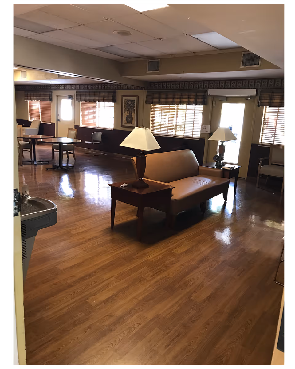 Interior view of a senior living facility common area with wooden flooring, a brown leather couch, two side tables each with a lamp, several chairs, round tables, windows with blinds, and a water fountain on the left side.