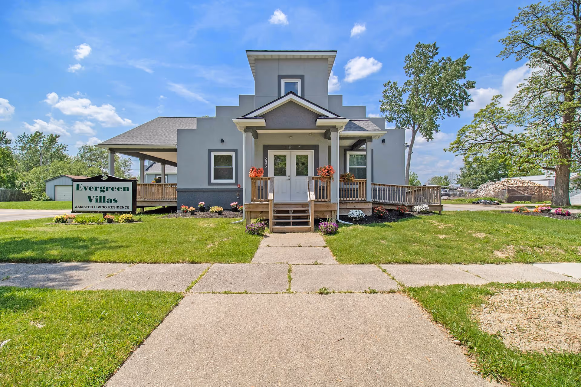 Front exterior view of a single-story assisted living residence named Evergreen Villas, featuring a gray building with a small porch, white double doors, flower pots, and a green lawn with a sidewalk leading to the entrance under a blue sky with scattered clouds.