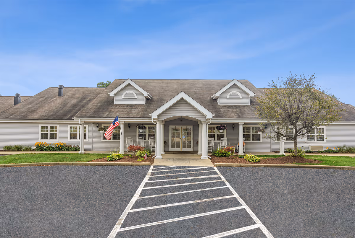 Front exterior view of a single-story senior living facility building with a covered entrance supported by white columns, an American flag, rocking chairs on the porch, landscaped flower beds, and a tree on the right side under a clear blue sky.