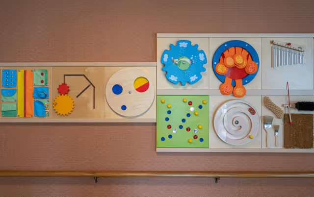 A wall-mounted sensory activity board featuring various colorful interactive elements including gears, buttons, a spinning disc, a xylophone, and textured brushes, designed for cognitive and motor skill engagement.