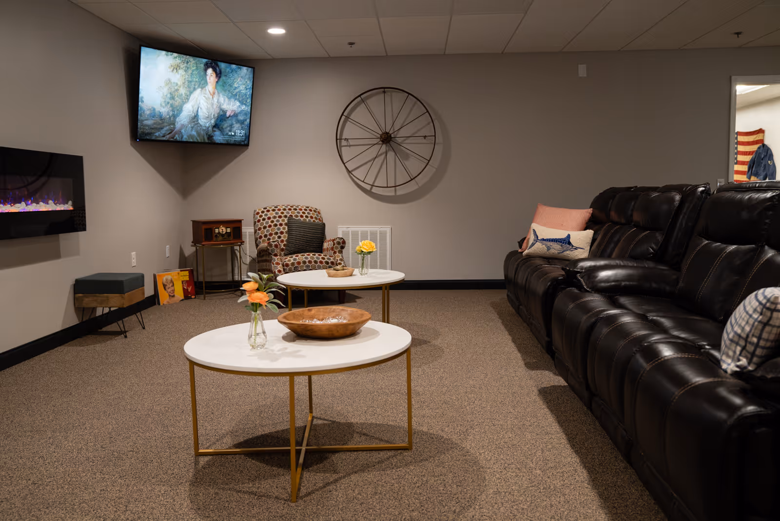 A cozy living room with a large black leather sofa adorned with pillows, two round white coffee tables with gold legs holding small flower vases and a wooden bowl, a patterned armchair with a pillow, a wall-mounted TV displaying a painting of a woman, a decorative large wheel on the wall, and a modern electric fireplace on the left wall.