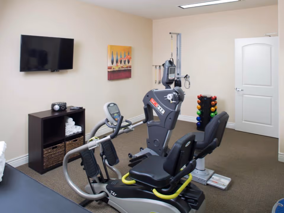 A small exercise room with a recumbent exercise bike, a rack of colorful dumbbells, a wall-mounted TV, a small shelf with towels and baskets, and a piece of wall art. The room has beige walls and carpeted floor.