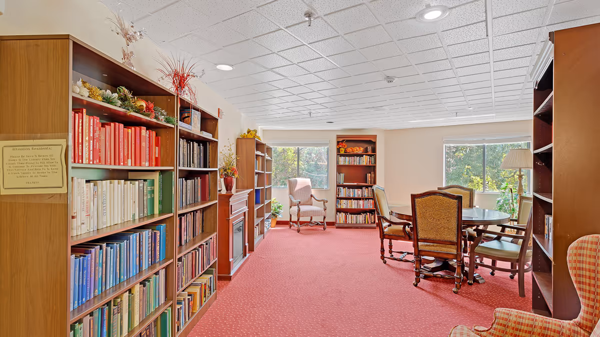 A cozy library room with bookshelves filled with books arranged by color, a fireplace, a round table with four upholstered chairs, and large windows letting in natural light. The room has a red carpet and decorative plants on top of the shelves.