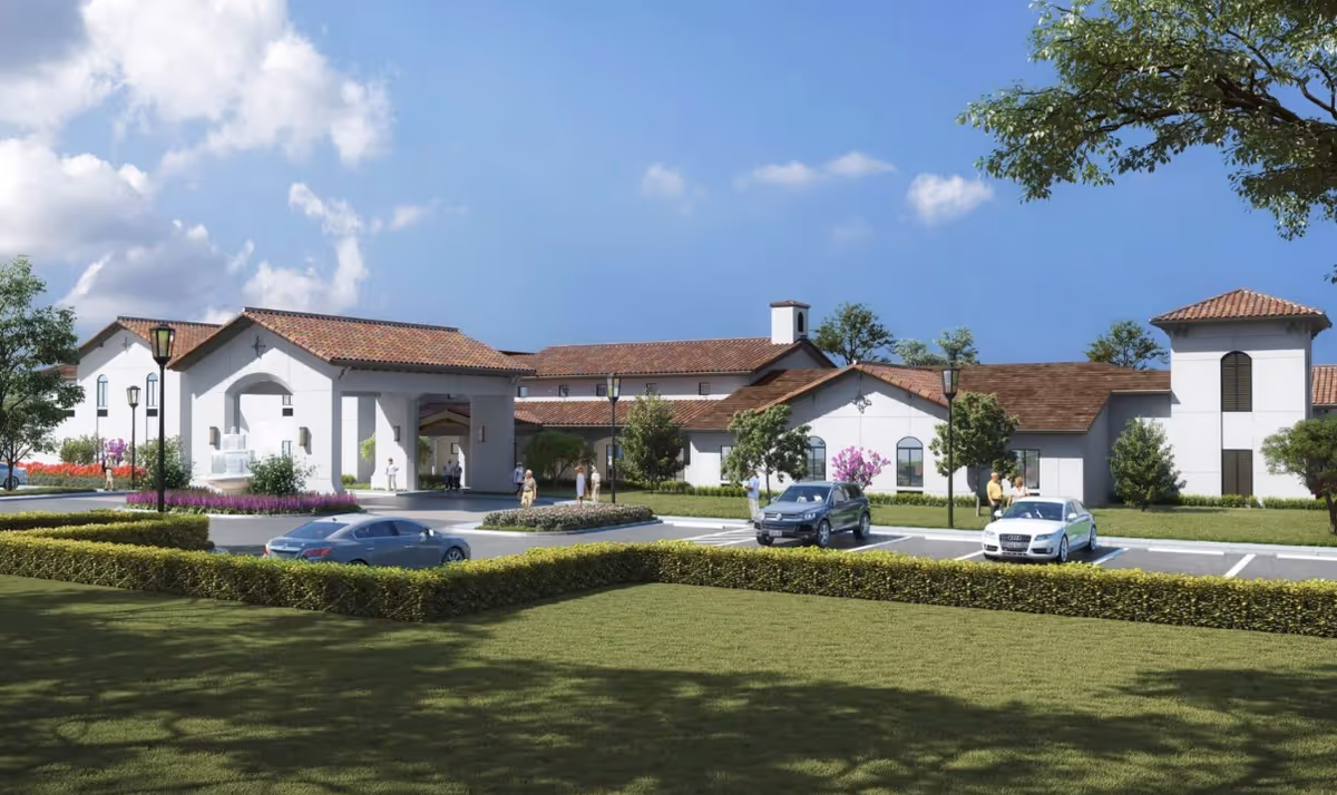 Exterior view of a senior living facility named HearthStone at Wildwood, featuring white buildings with red-tiled roofs, a parking lot with several cars, landscaped greenery, and a few people walking near the entrance under a partly cloudy sky.
