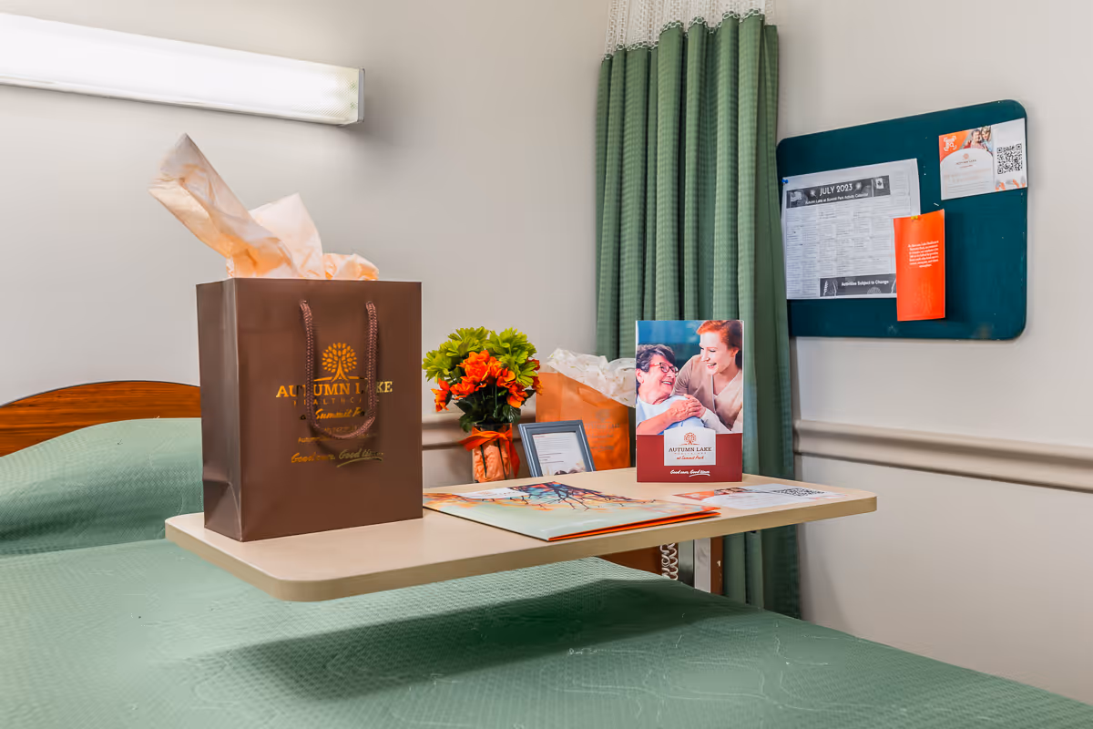 A hospital or healthcare facility room with a bed covered in a green blanket. A small table over the bed holds a brown gift bag with tissue paper, a small bouquet of orange flowers, a framed photo, and a brochure featuring two smiling women. A green privacy curtain and a bulletin board with papers are visible in the background.