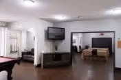 Interior view of a common area in a retirement center featuring a wall-mounted flat-screen TV above a cabinet, a pool table partially visible on the left, and a dining area with a table and chairs in the background. The room has white walls, dark wood flooring, and ceiling lights.