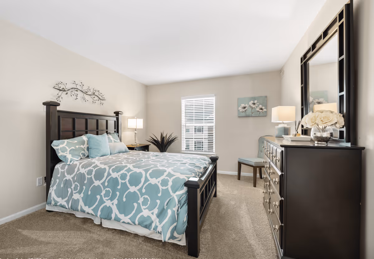 A bright bedroom with a large dark wooden bed frame and a blue and white patterned bedspread. There are two pillows on the bed, a nightstand with a lamp on the left side, a window with blinds in the center, and a dark wooden dresser with a large mirror on the right. A chair and a small piece of wall art with flowers are also visible.